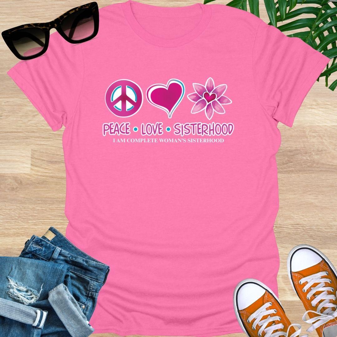 Pink t-shirt with 'Peace Love Sisterhood' design on a wooden surface with sunglasses, jeans, and sneakers.