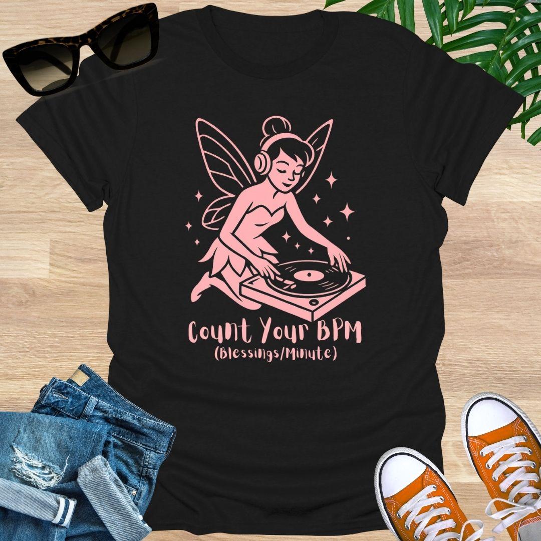 Black t-shirt with a pink fairy DJ graphic and text on a wooden surface with sunglasses, jeans, and sneakers.
