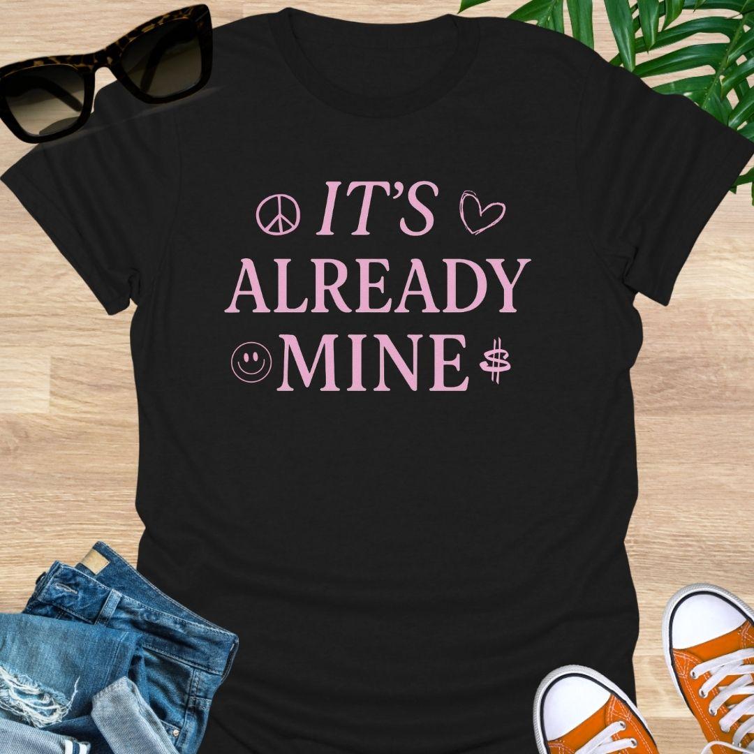 Pink text “It’s Already Mine” with peace, heart, smiley, and dollar icons on a black manifestation graphic tee.