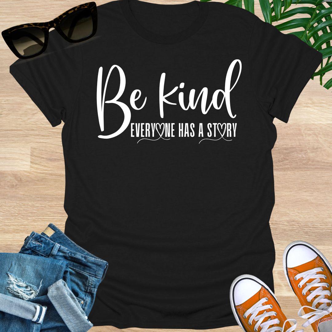 “Be Kind—Everyone Has a Story” in white script with heart loops on a black unisex tee.