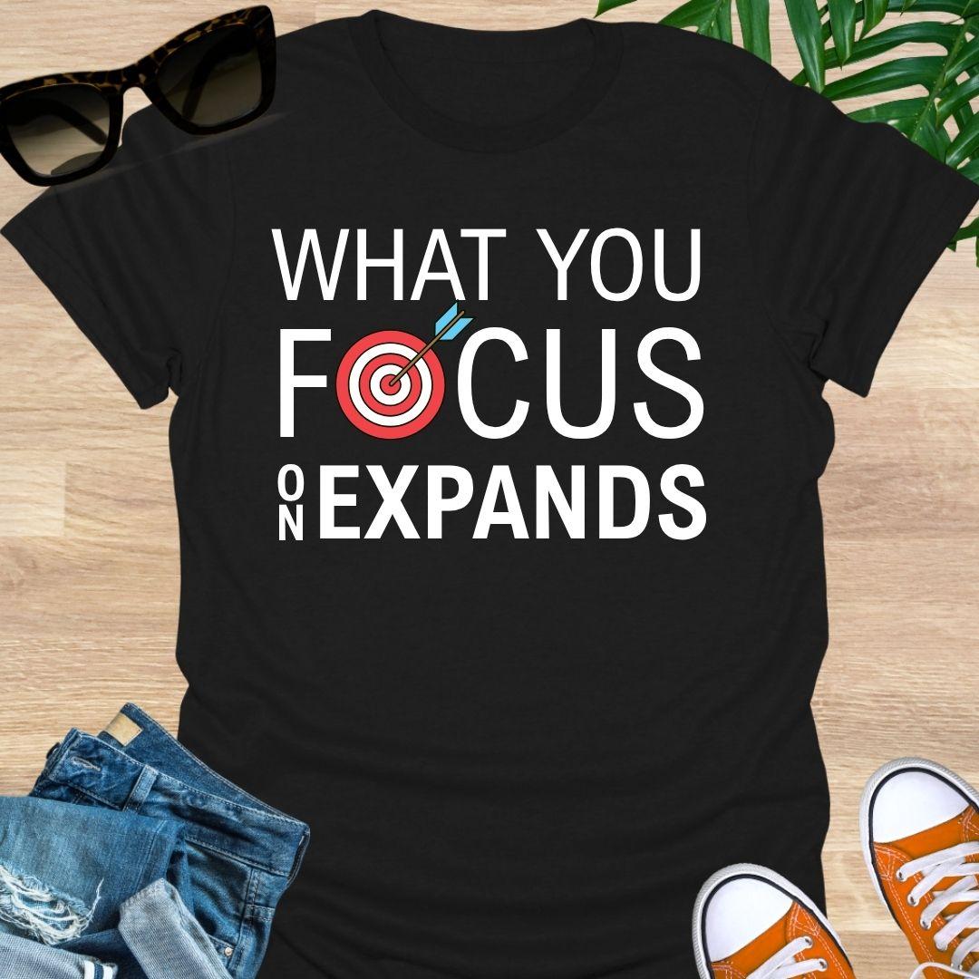 “What You Focus On Expands” text with red bullseye and arrow on a black unisex tee.