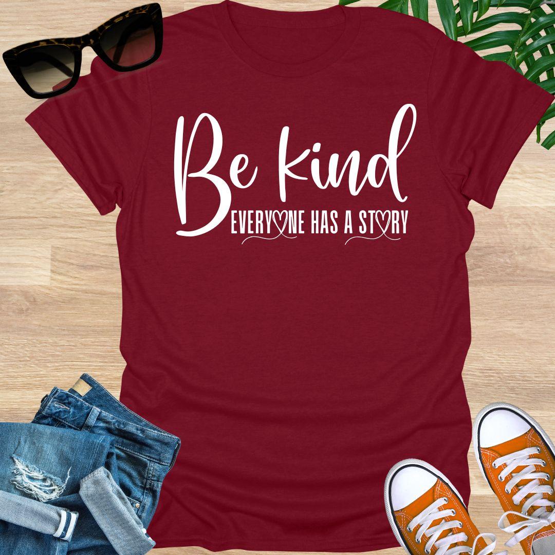 “Be Kind—Everyone Has a Story” in white script with heart loops on a red unisex tee.