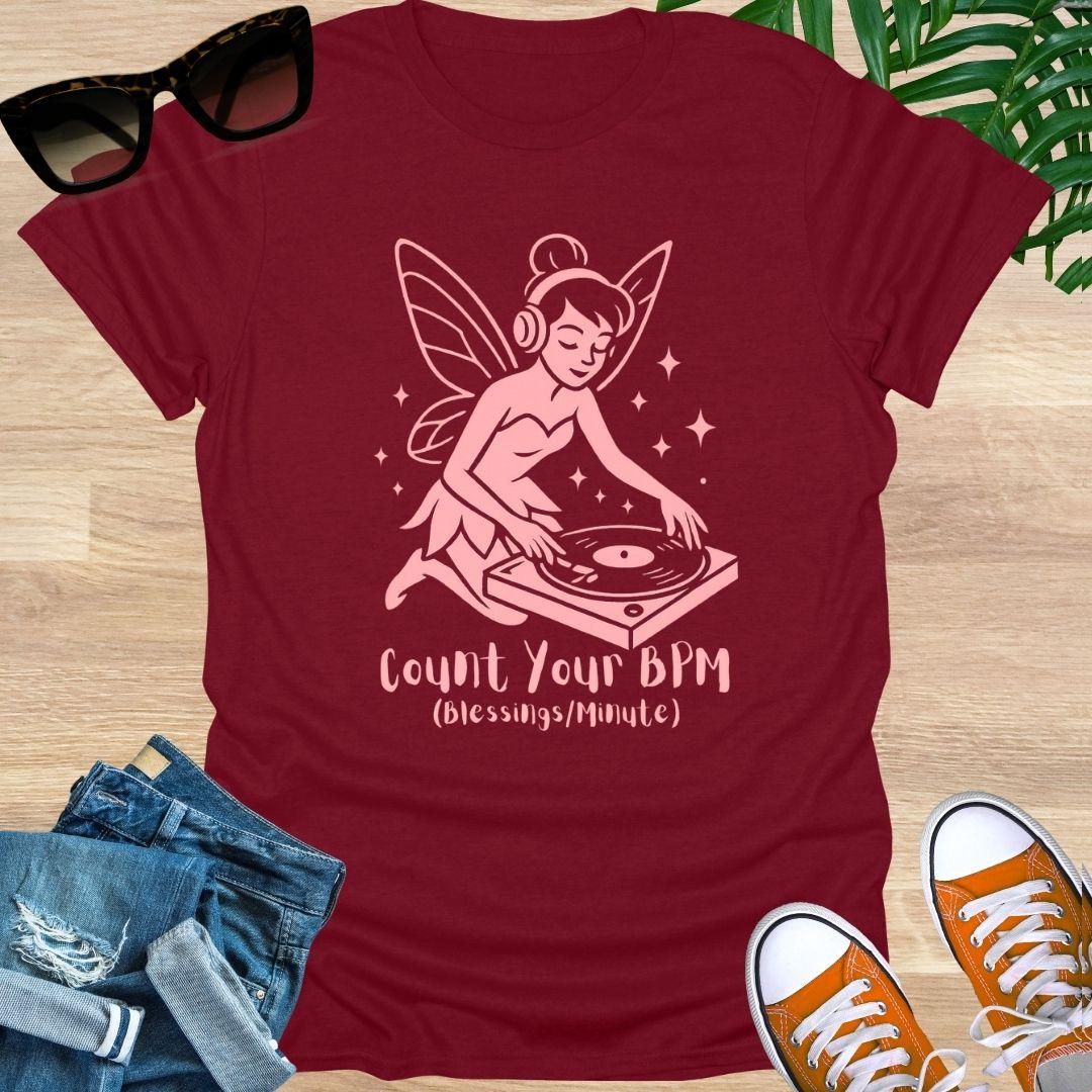 Red t-shirt with a graphic of a fairy on a turntable and text 'Count Your BPM (Blessings Per Minute)' on a wooden surface with sunglasses, jeans, and sneakers.