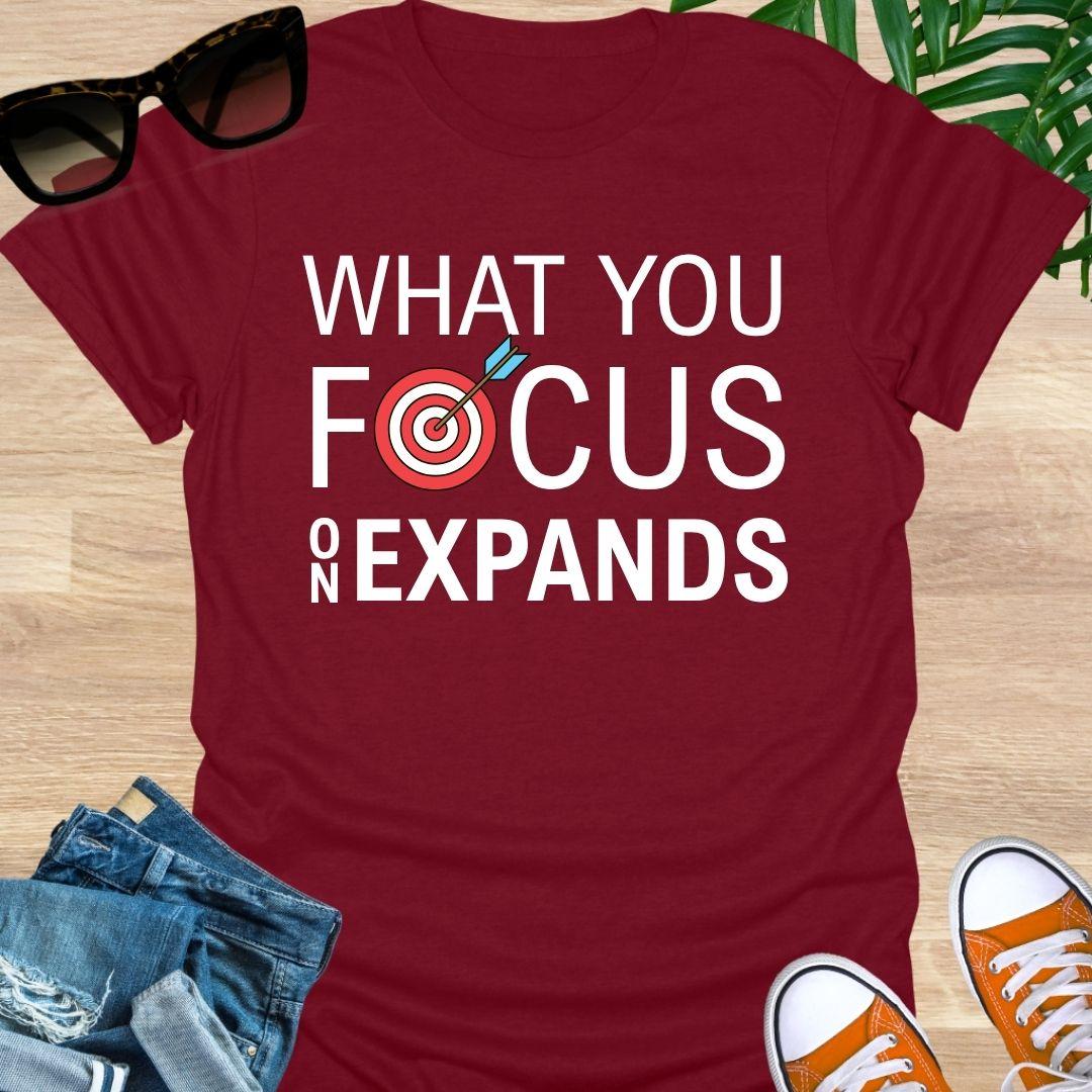 “What You Focus On Expands” text with red bullseye and arrow on a red unisex tee.