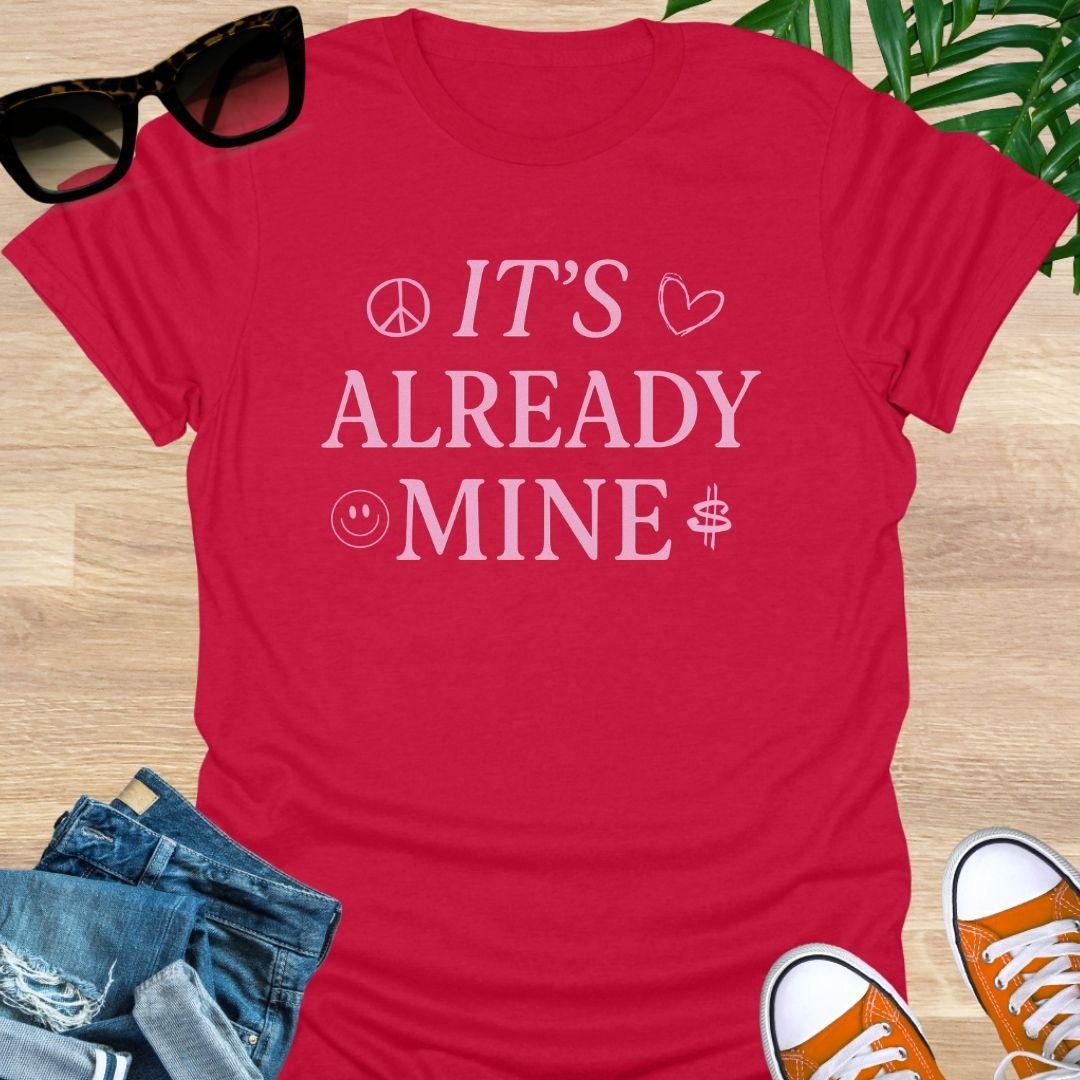 Pink text “It’s Already Mine” with peace, heart, smiley, and dollar icons on a red manifestation graphic tee.