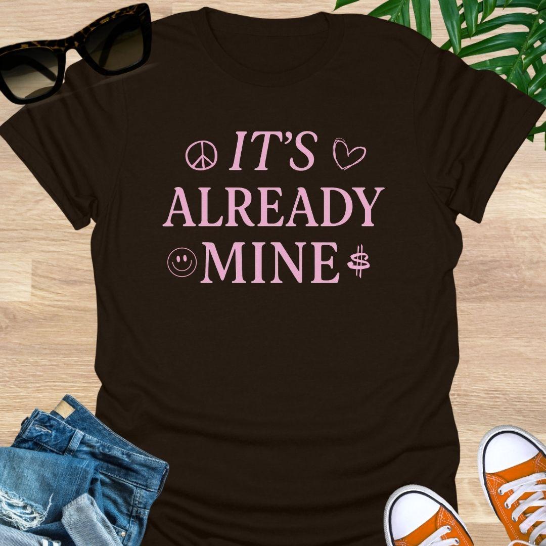 Pink text “It’s Already Mine” with peace, heart, smiley, and dollar icons on a brown manifestation graphic tee.