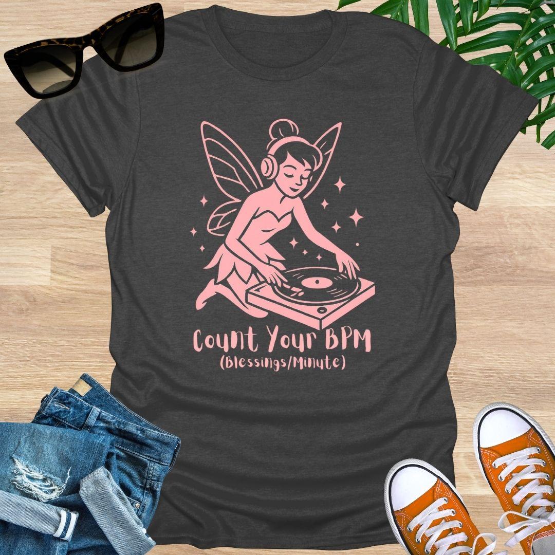 Black t-shirt with pink fairy graphic and text 'Count Your BPM' on a wooden surface with sunglasses, jeans, and sneakers.