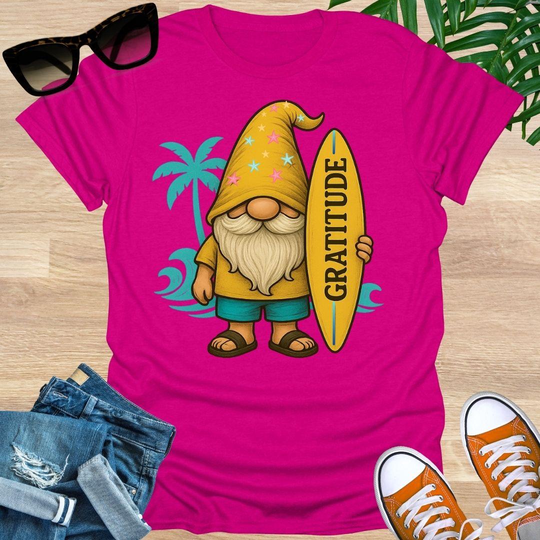 Pink t-shirt with a gnome holding a 'Gratitude' surfboard graphic, sunglasses, jeans, and shoes on a wooden surface.