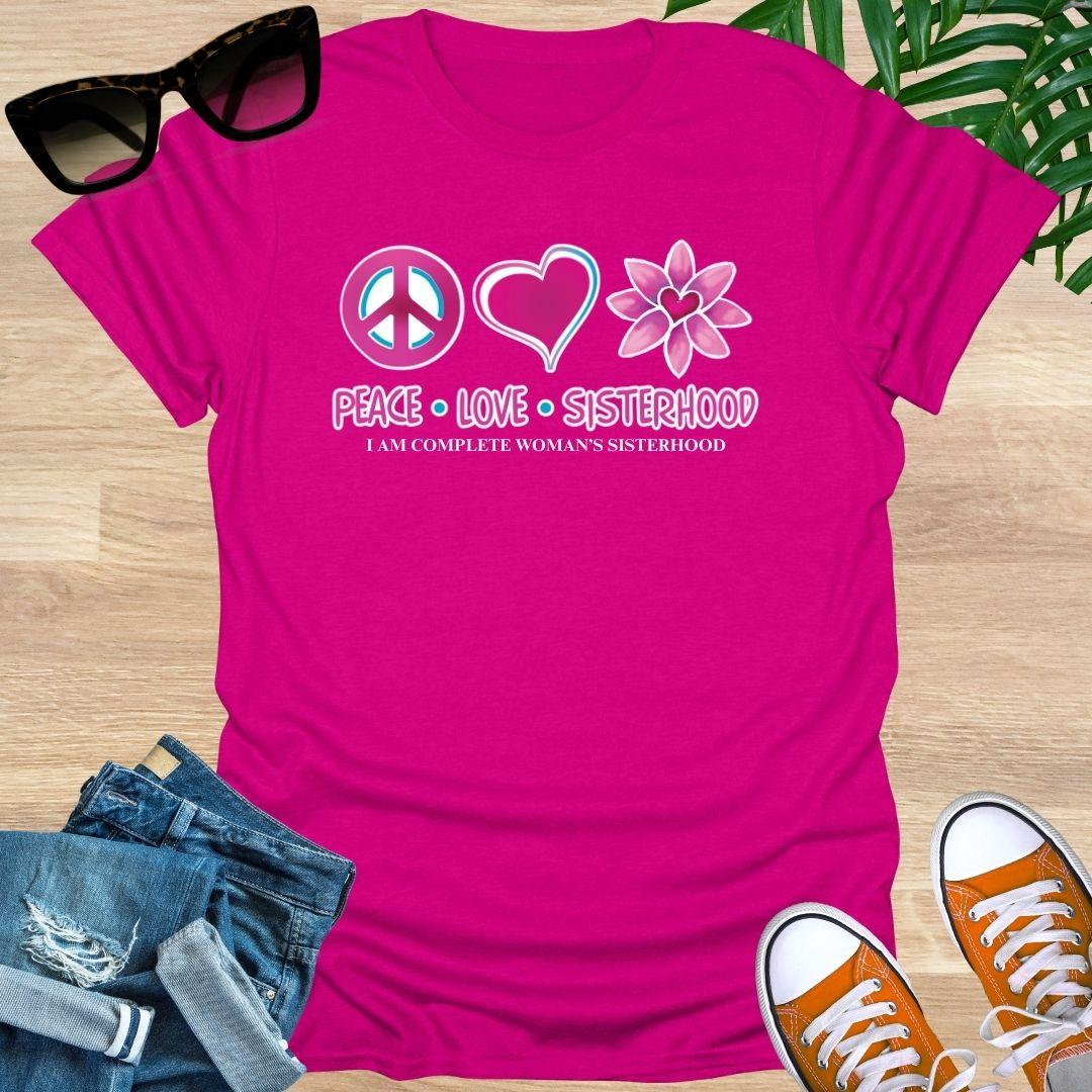 Pink t-shirt with 'Peace Love Sisterhood' design on a wooden surface with sunglasses, jeans, and sneakers.