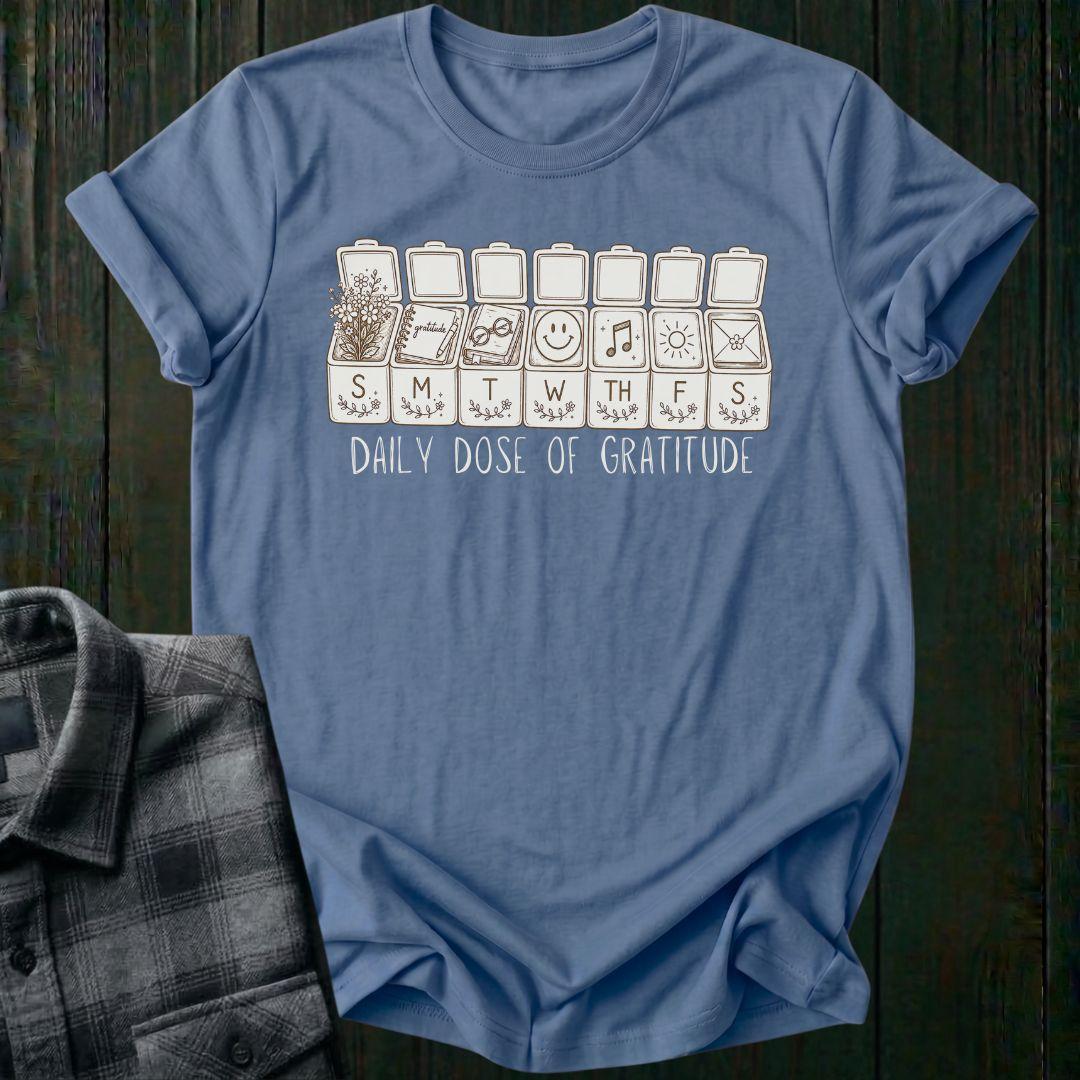 Vintage-inspired weekly pill organizer graphic featuring gratitude-themed icons for each day of the week, including flowers, journaling, reading, smiling, music, sunshine, and connection, with the phrase Daily Dose of Gratitude on a indigo blue tee
