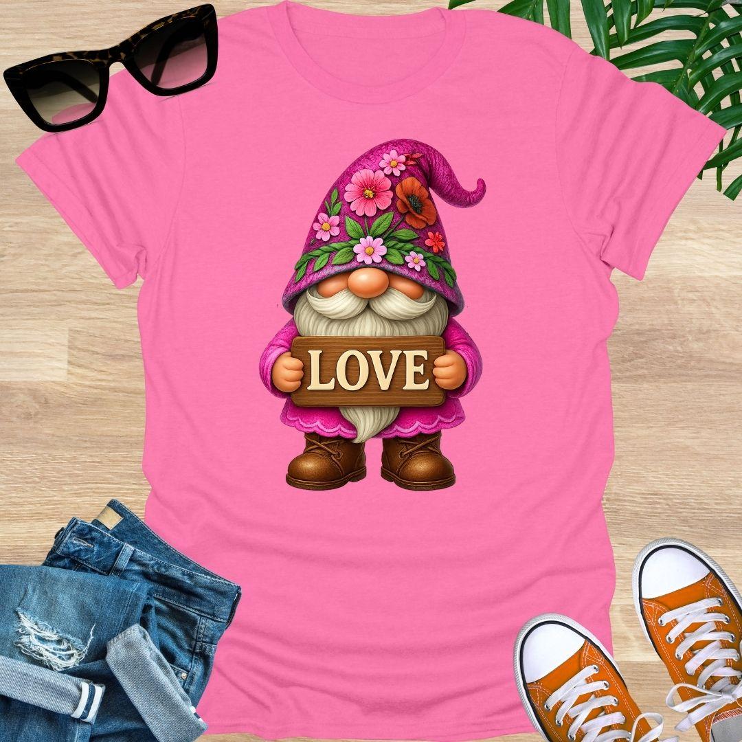 Pink t-shirt with a gnome holding a 'LOVE' sign, surrounded by sunglasses, jeans, and shoes on a wooden surface.