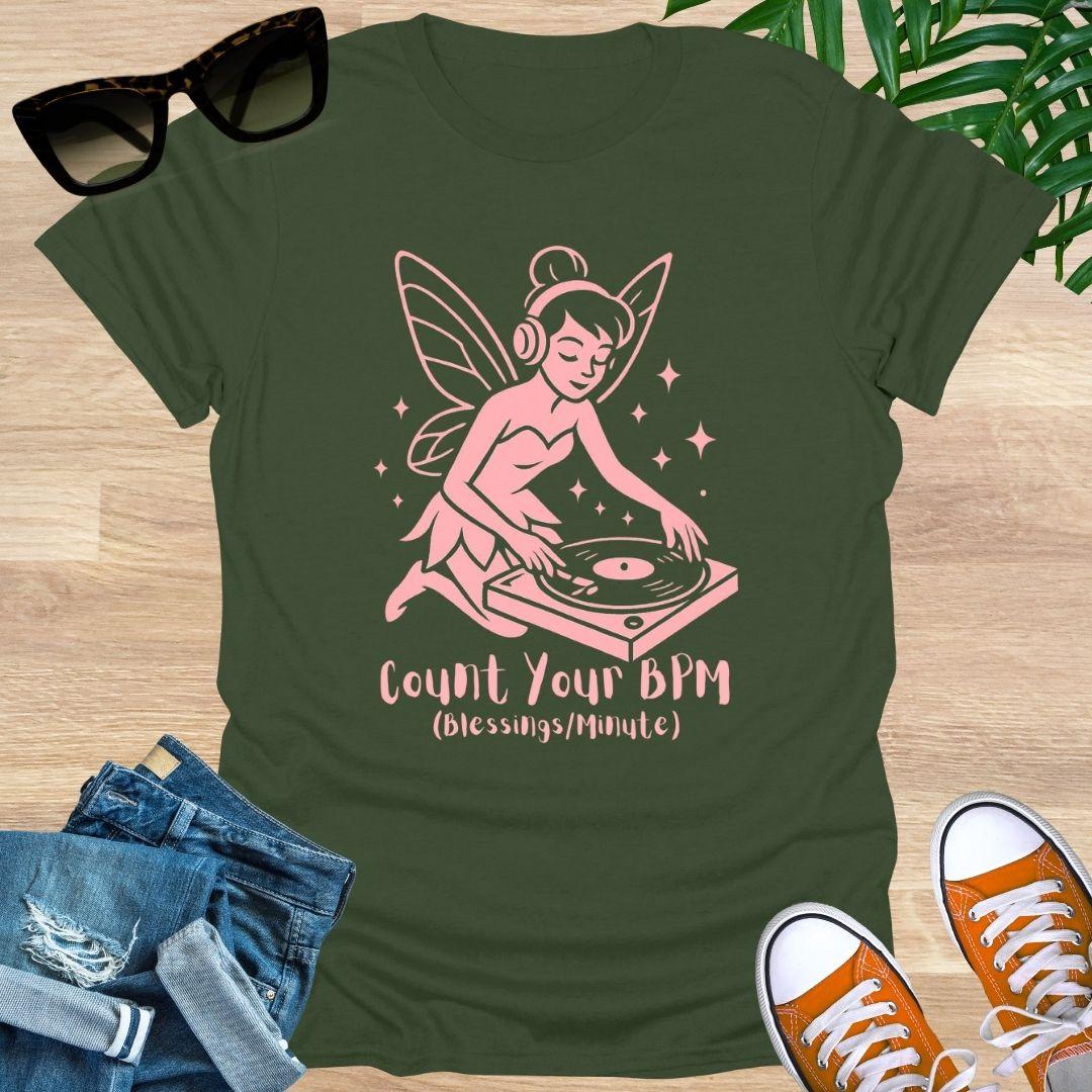 Green t-shirt with a pink fairy DJ design and text 'Count Your BPM' on a wooden surface with sunglasses, jeans, and sneakers.