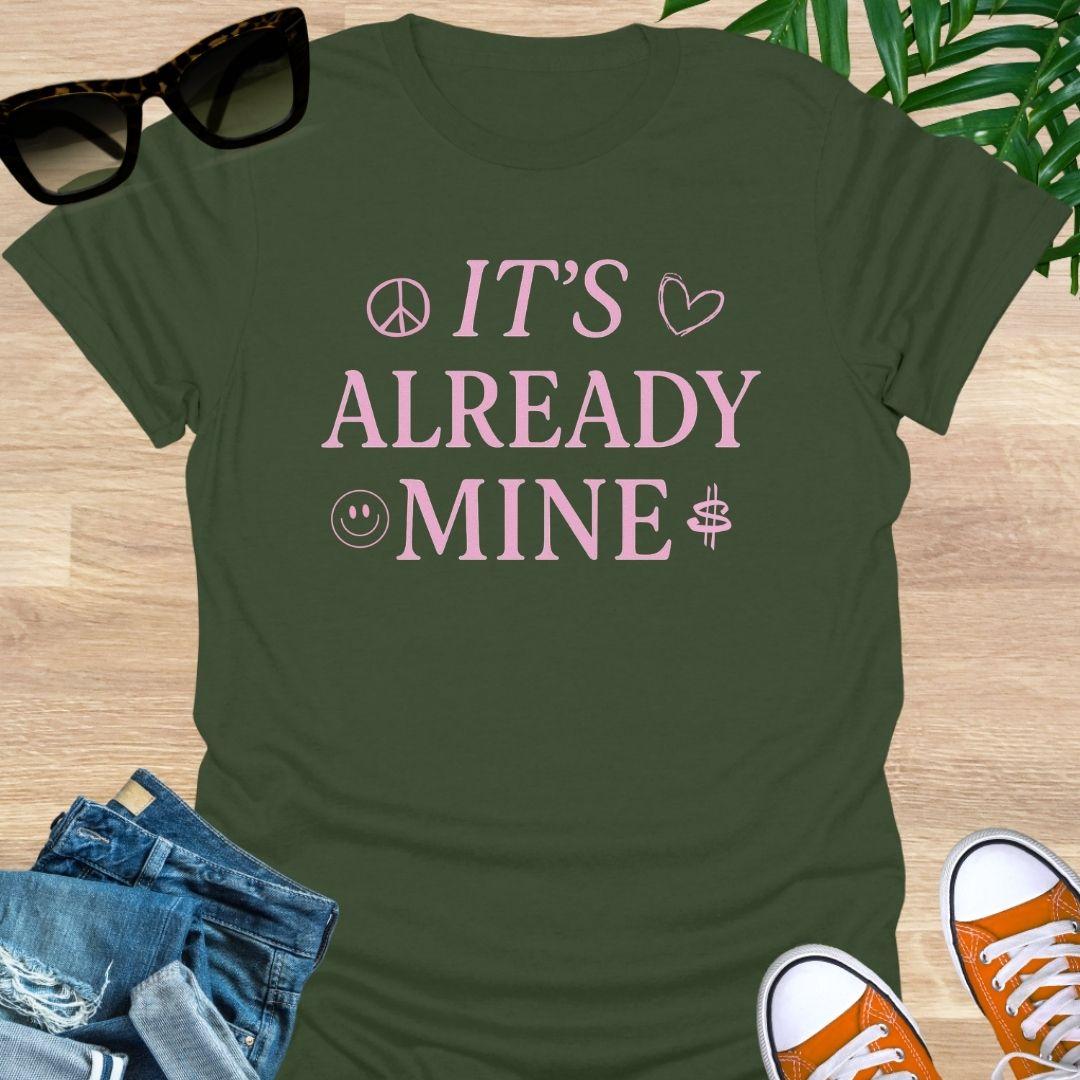 Pink text “It’s Already Mine” with peace, heart, smiley, and dollar icons on a military green manifestation graphic tee.