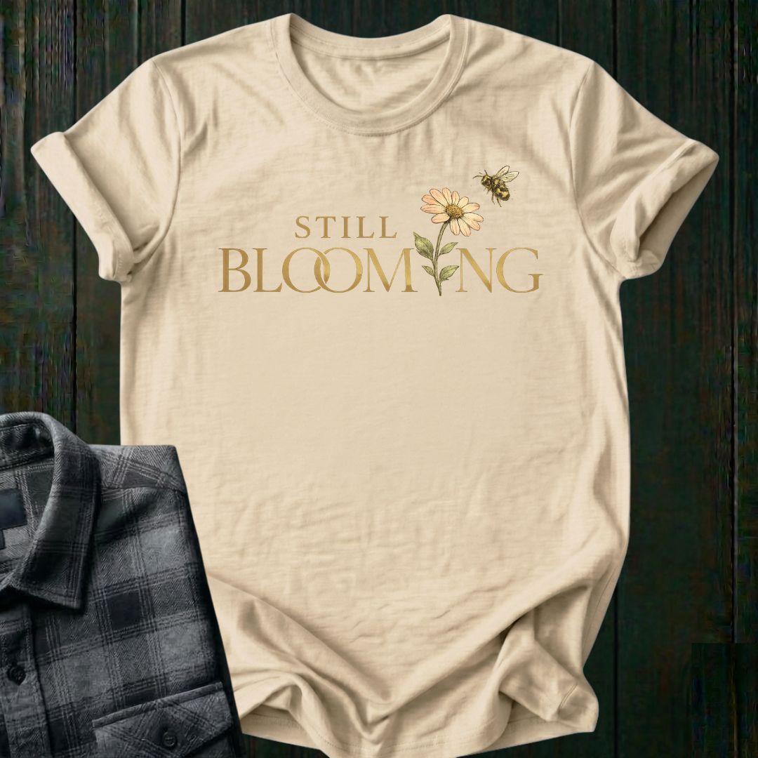Elegant typography design featuring the phrase "Still Blooming" with a blooming flower growing through the lettering and a bee flying overhead on a beige shirt