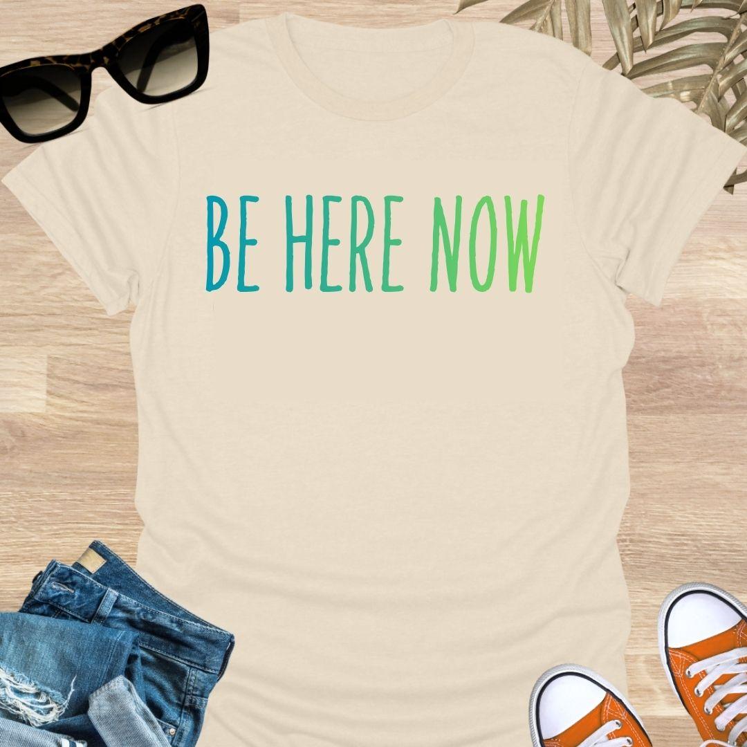 Blue-green gradient “Be Here Now” text on beige unisex mindfulness tee; reminder to breathe and be present.