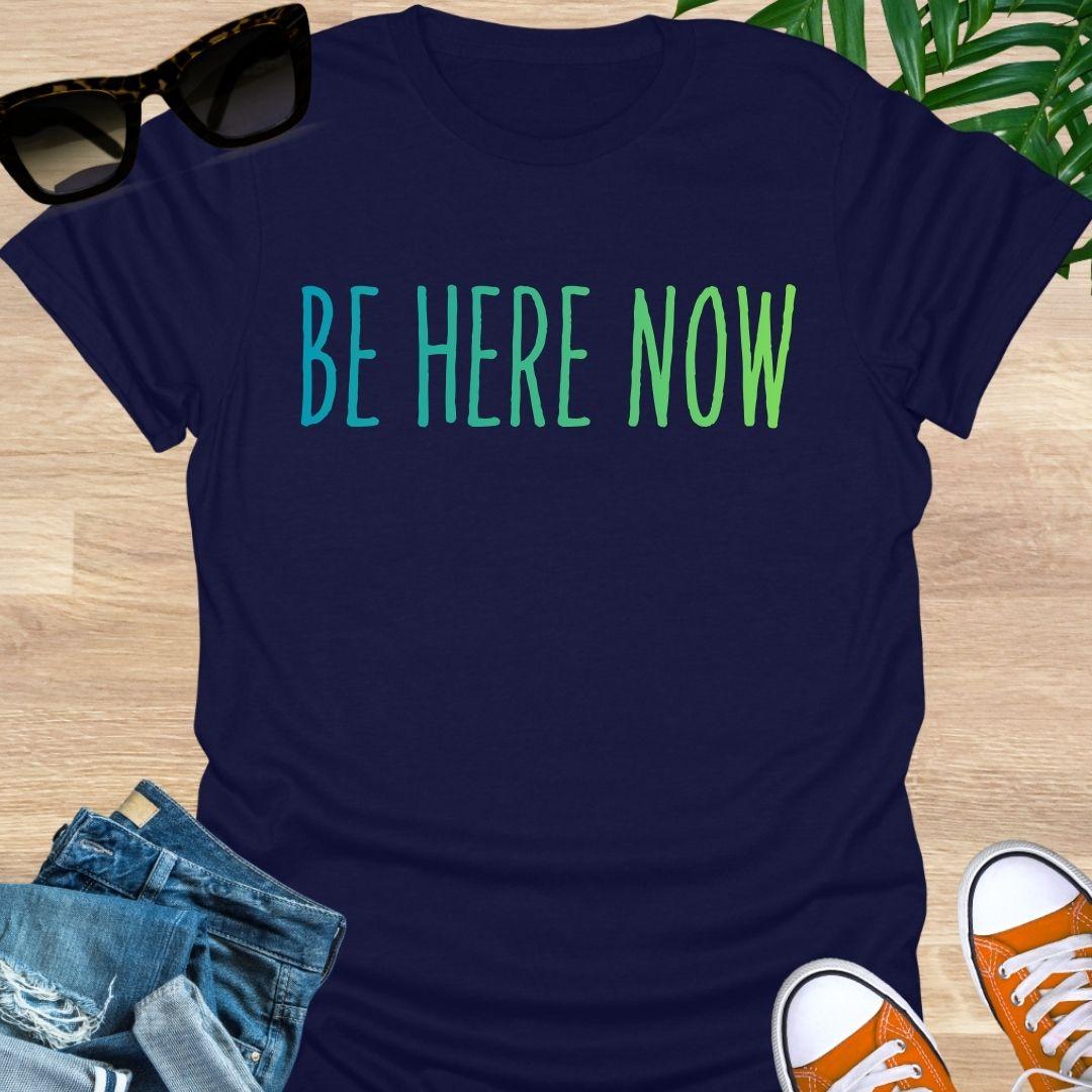 Blue-green gradient “Be Here Now” text on navy unisex mindfulness tee; reminder to breathe and be present.