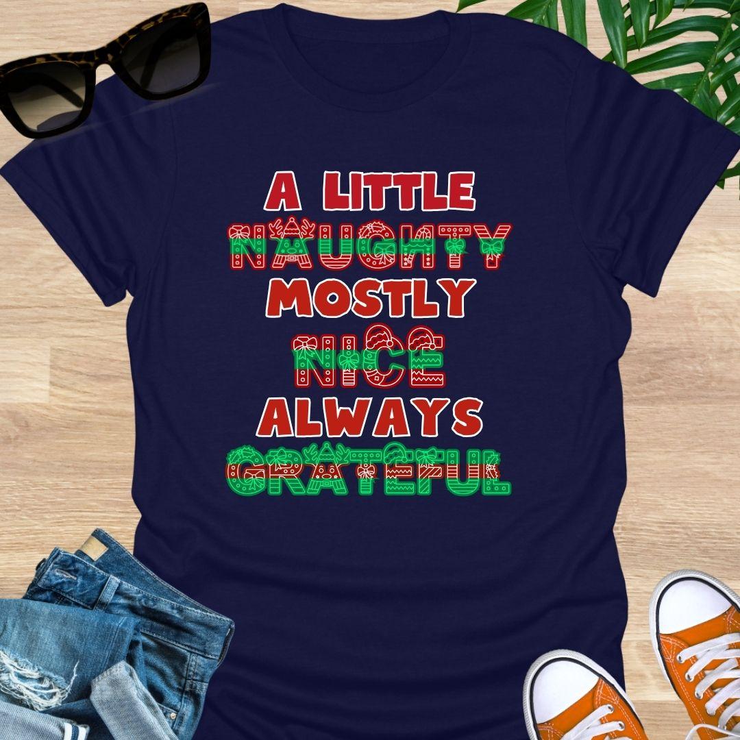 Red-green holiday letters spelling “A Little Naughty, Mostly Nice, Always Grateful” with bows, wreaths, and reindeer on a navy unisex tee.