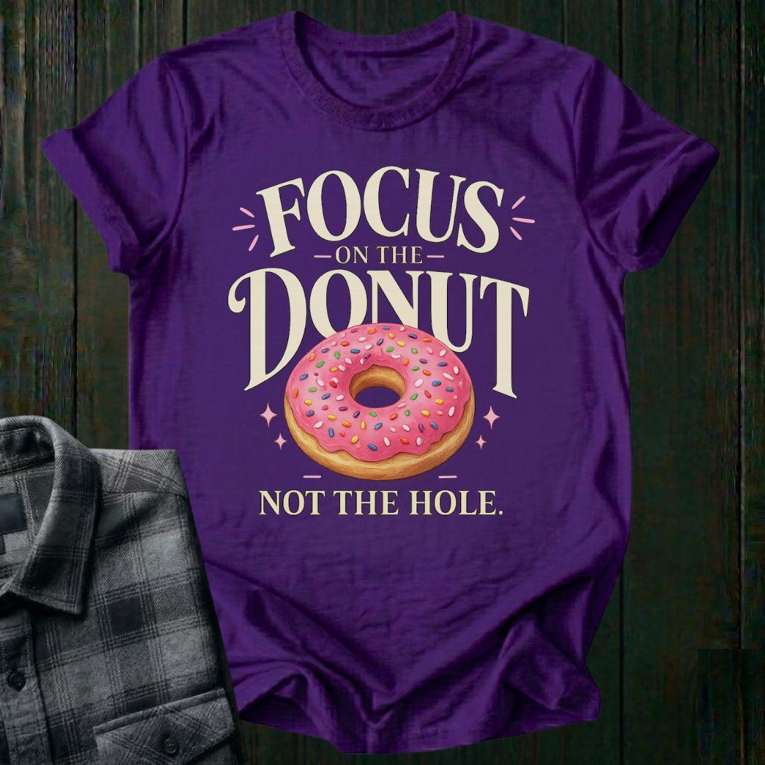 Typography design featuring the phrase "Focus on the Donut, Not the Hole" with a pink sprinkled donut, symbolizing gratitude, perspective, and focusing on life's blessings on a purple tee