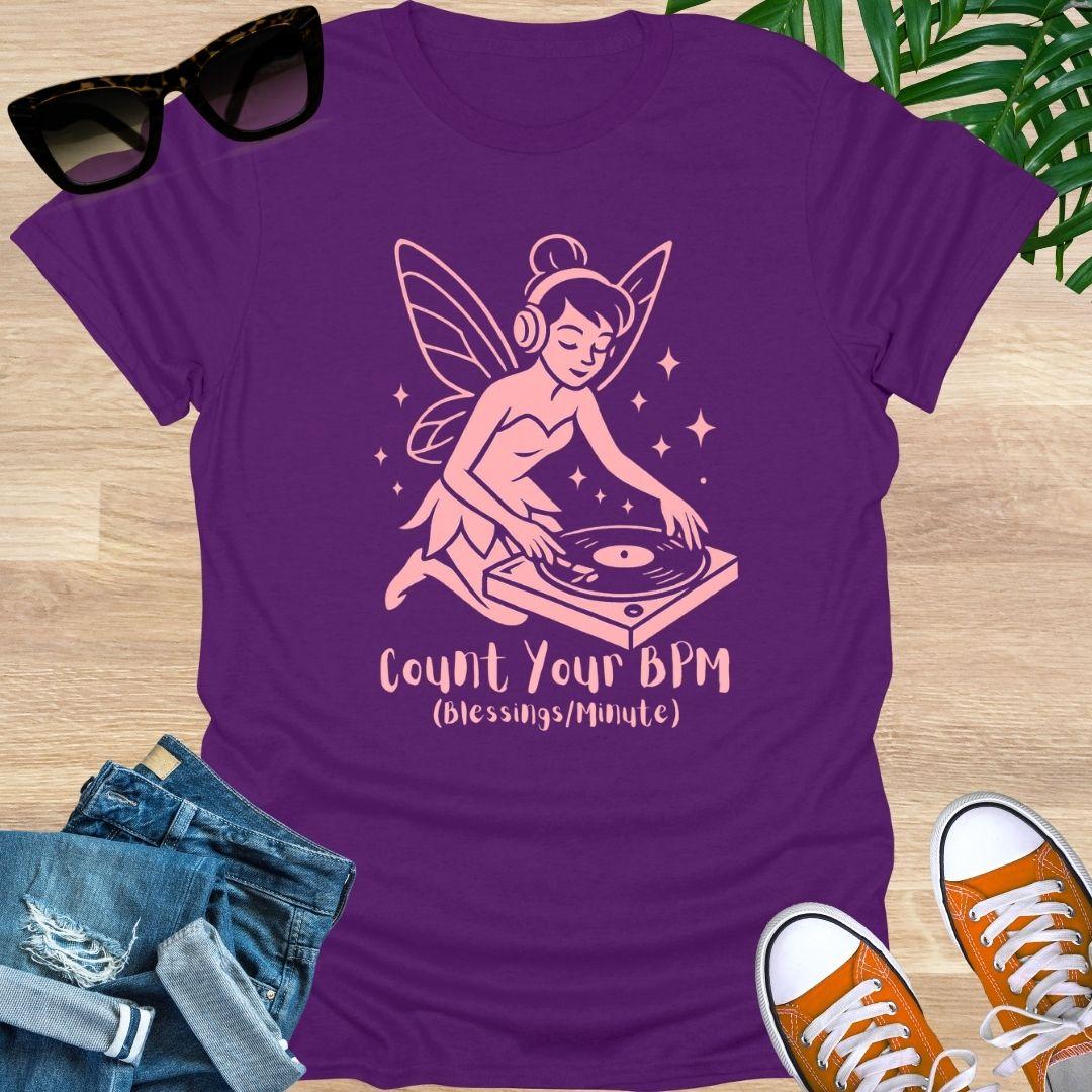 Purple t-shirt with a fairy DJ graphic and text on a wooden surface with sunglasses, jeans, and sneakers.