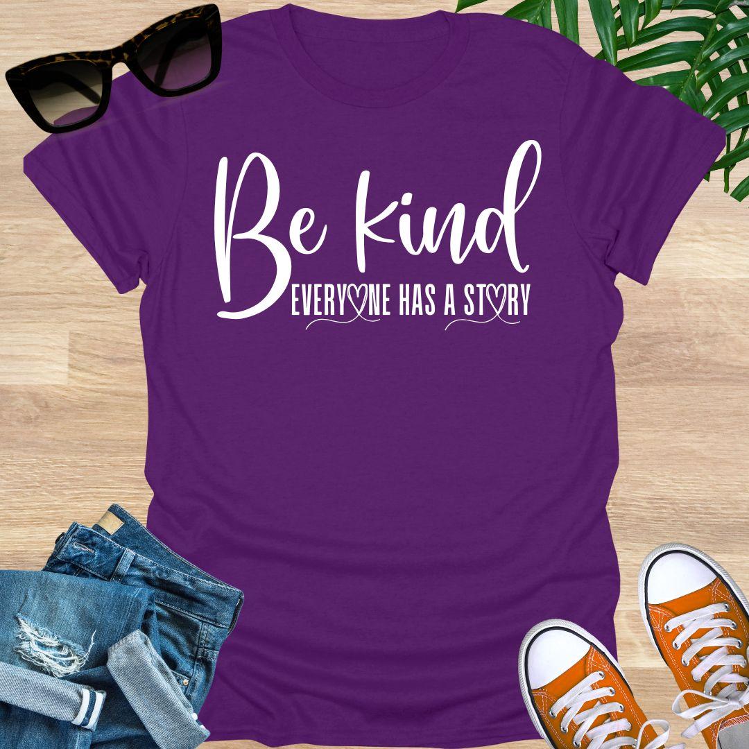 “Be Kind—Everyone Has a Story” in white script with heart loops on a purple unisex tee.