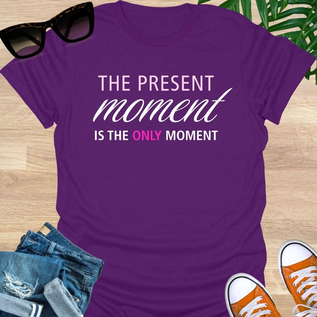 Purple tee with pink/white text reading “The Present Moment Is the Only Moment” minimalist mindfulness graphic.