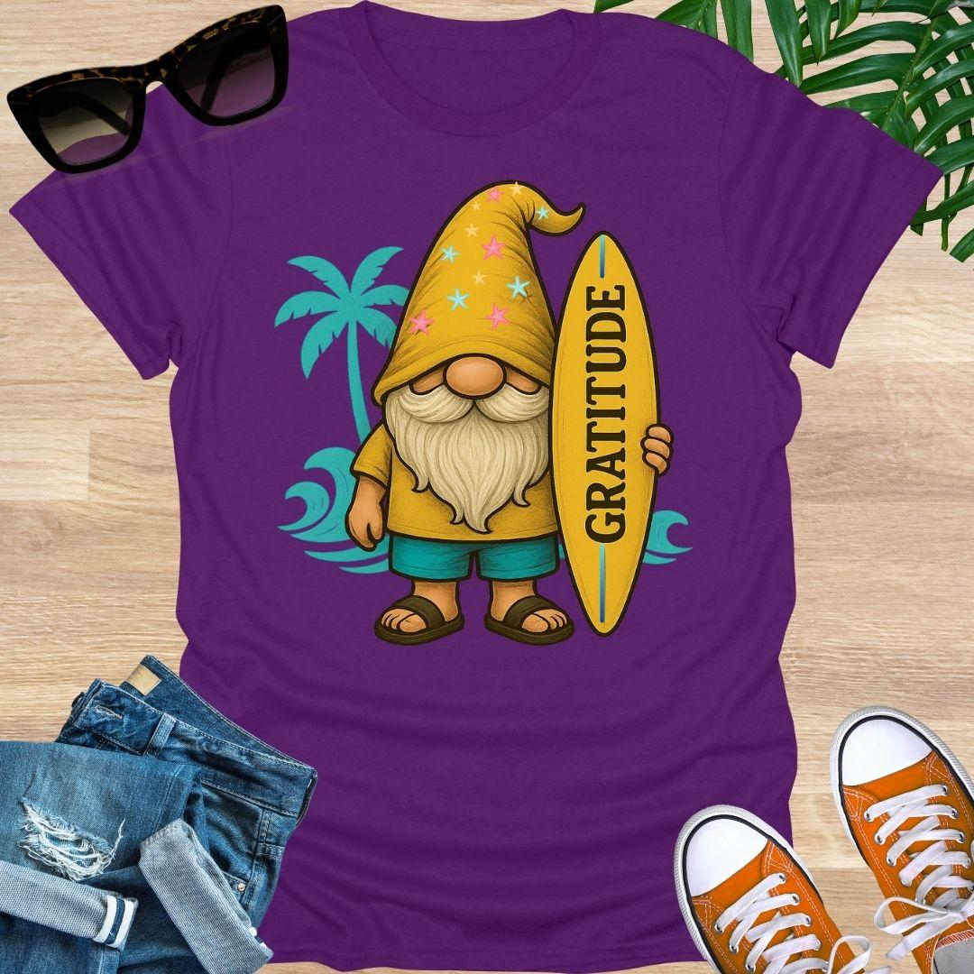 Purple t-shirt with a gnome holding a 'Gratitude' surfboard design, sunglasses, jeans, and sneakers on a wooden surface.