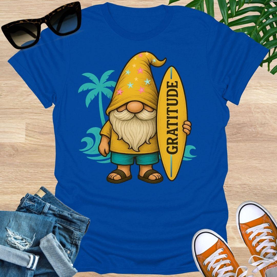 Blue t-shirt with a cartoon gnome holding a surfboard with 'Gratitude' on it, sunglasses, jeans, and shoes on a wooden surface.
