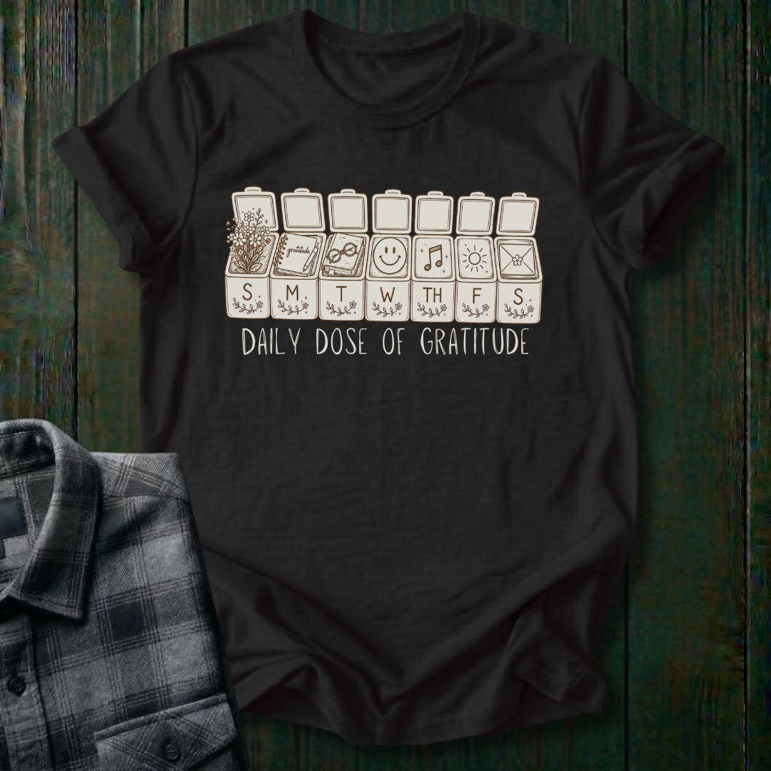 Vintage-inspired weekly pill organizer graphic featuring gratitude-themed icons for each day of the week, including flowers, journaling, reading, smiling, music, sunshine, and connection, with the phrase Daily Dose of Gratitude on a black tee