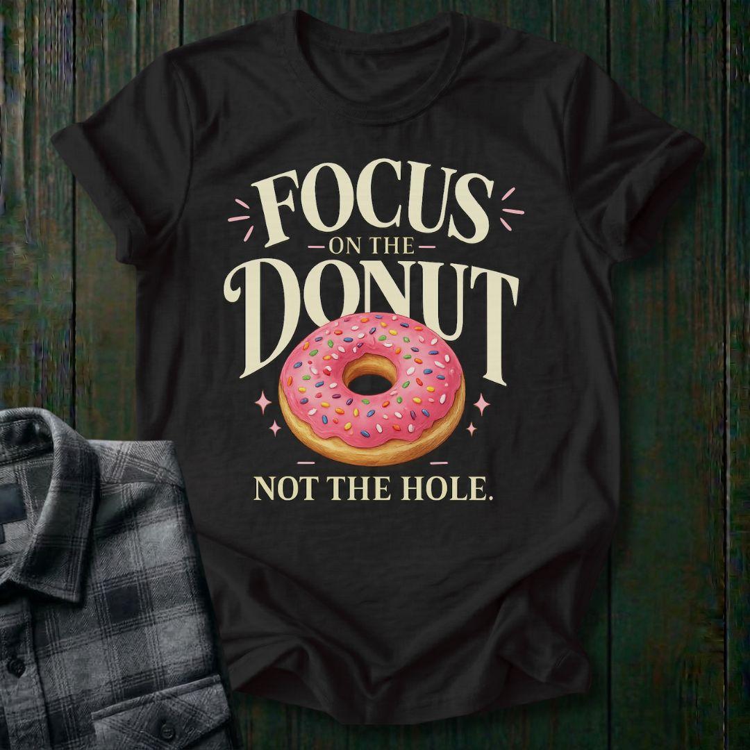 Typography design featuring the phrase "Focus on the Donut, Not the Hole" with a pink sprinkled donut, symbolizing gratitude, perspective, and focusing on life's blessings on a black tee