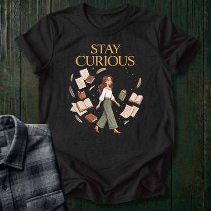 Illustrated woman walking among floating books, journals, feathers, and stars beneath the words "Stay Curious," symbolizing lifelong learning, creativity, and wonder on a black tee.