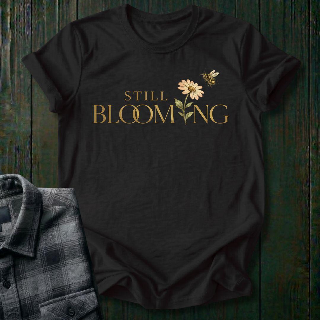 Elegant typography design featuring the phrase "Still Blooming" with a blooming flower growing through the lettering and a bee flying overhead on a black shirt