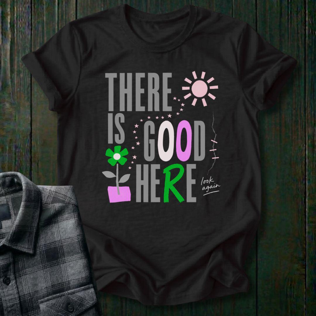 Colorful typography design featuring the phrase "There Is Good Here" with flowers, stars, sunshine, and the handwritten words "look again" on a black tee.