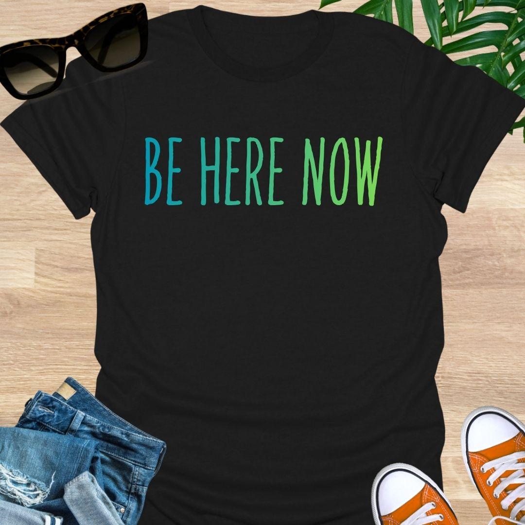 Blue-green gradient “Be Here Now” text on black unisex mindfulness tee; reminder to breathe and be present.