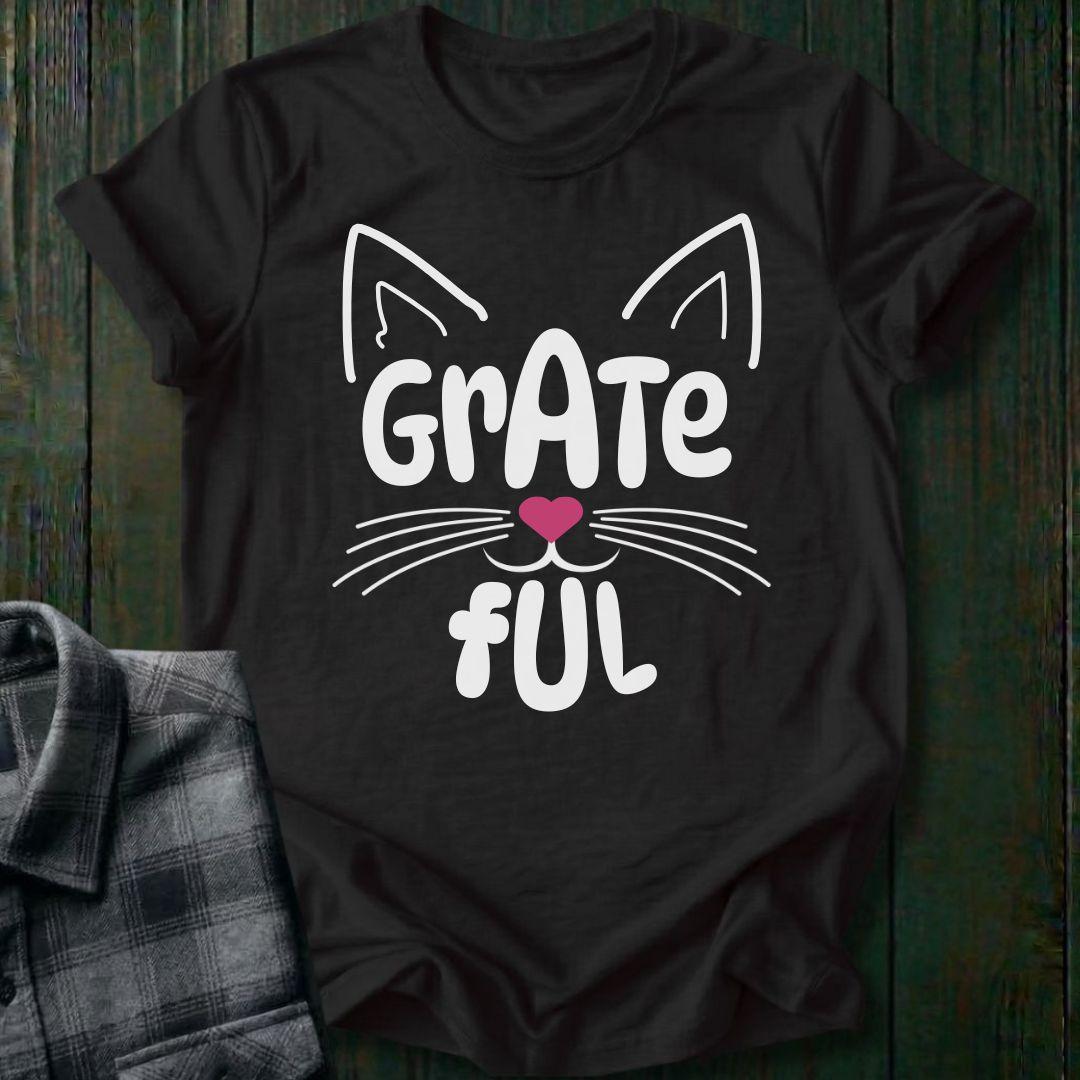 Black t-shirt with a cat face design and 'Grate Ful' text on a wooden surface.
