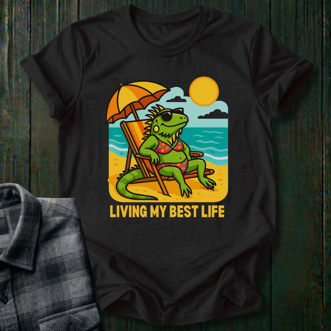 Black t-shirt with a graphic of a lizard on a beach and 'Living My Best Life' text, placed on a wooden surface.