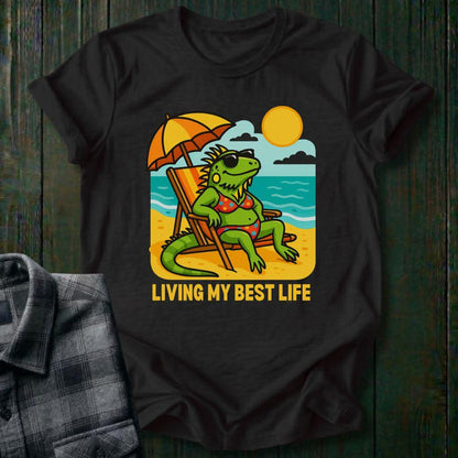 Black t-shirt with a graphic of a lizard on a beach and 'Living My Best Life' text, placed on a wooden surface.
