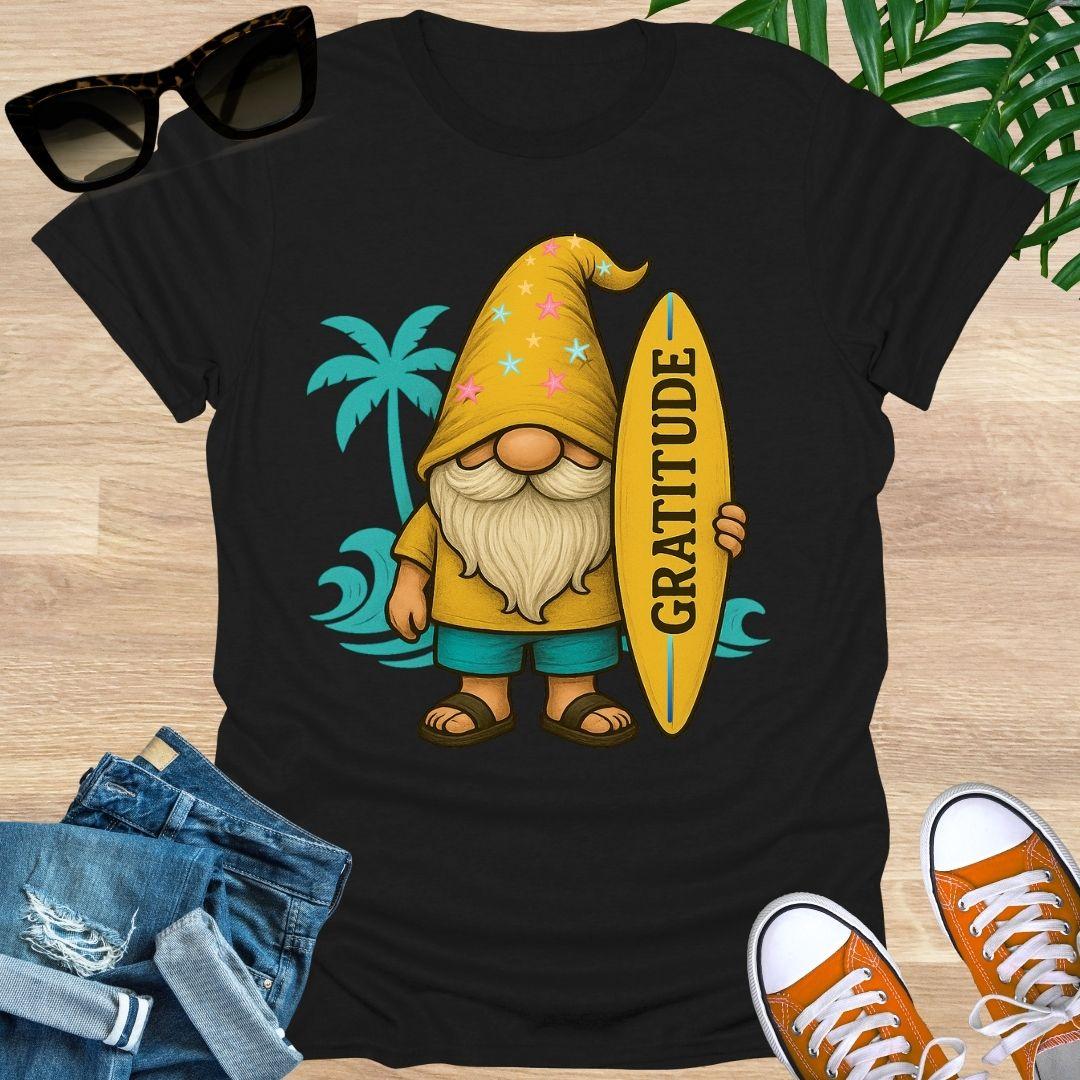 Black t-shirt with a gnome holding a 'Gratitude' surfboard graphic, sunglasses, jeans, and sneakers on a wooden surface.