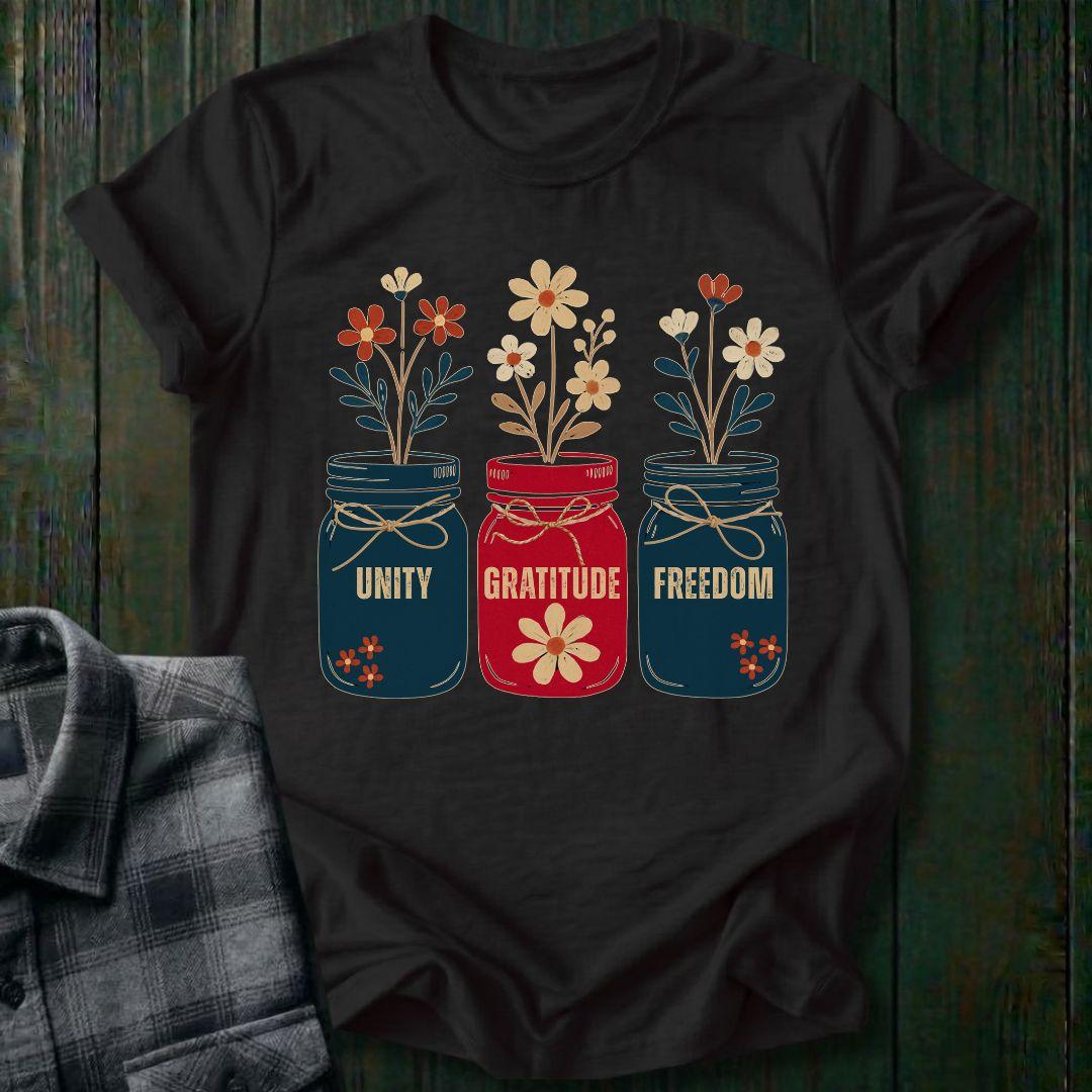 Black t-shirt with floral jars and text 'Unity', 'Gratitude', 'Freedom' on a wooden surface.