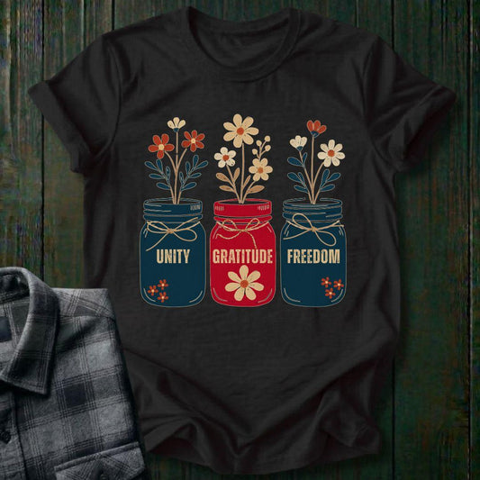 Black t-shirt with floral jars and text 'Unity', 'Gratitude', 'Freedom' on a wooden surface.