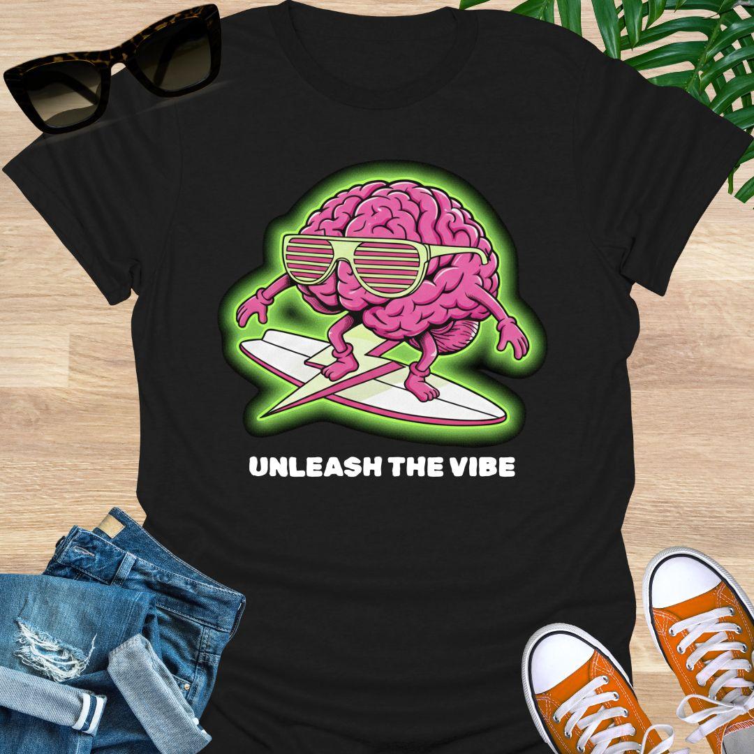 Pink cartoon brain wearing sunglasses surfing on a board with green glow and the words “Unleash the Vibe” on a black t-shirt