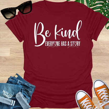 “Be Kind—Everyone Has a Story” in white script with heart loops on a red unisex tee.