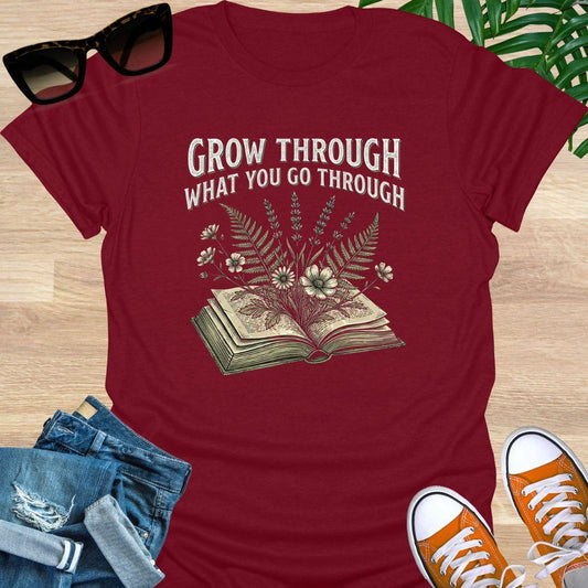 Vintage-style illustration of flowers growing from an open book with the phrase “Grow Through What You Go Through” on a red unisex t-shirt