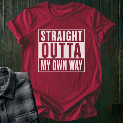 Red t-shirt with 'Straight Outta My Own Way' text on a wooden surface with a plaid shirt.