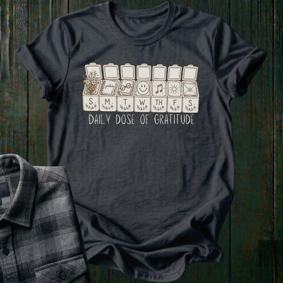 Vintage-inspired weekly pill organizer graphic featuring gratitude-themed icons for each day of the week, including flowers, journaling, reading, smiling, music, sunshine, and connection, with the phrase Daily Dose of Gratitude on a charcoal tee