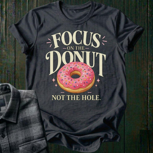 Typography design featuring the phrase "Focus on the Donut, Not the Hole" with a pink sprinkled donut, symbolizing gratitude, perspective, and focusing on life's blessings on a charcoal tee