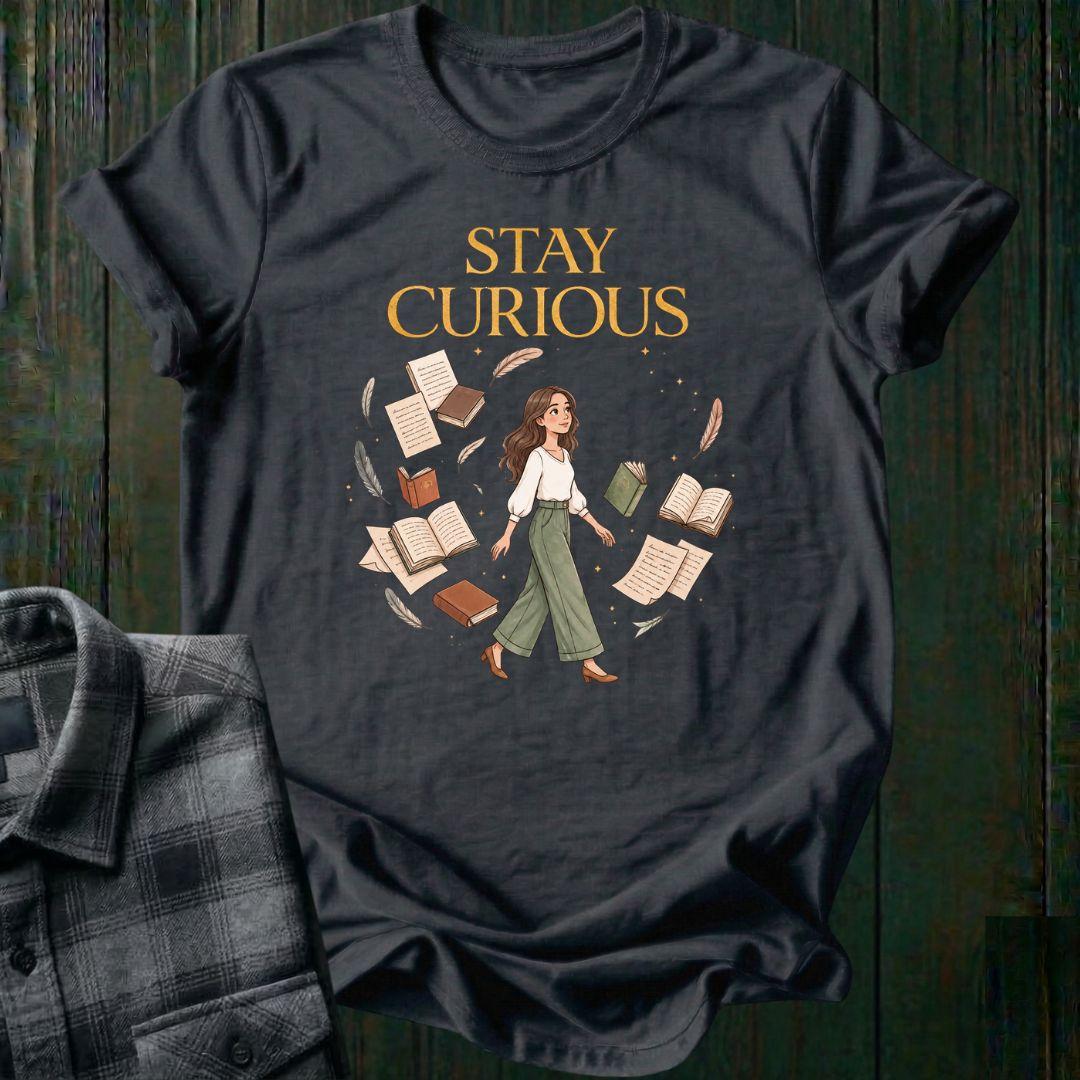 Illustrated woman walking among floating books, journals, feathers, and stars beneath the words "Stay Curious," symbolizing lifelong learning, creativity, and wonder on a charcoal tee.