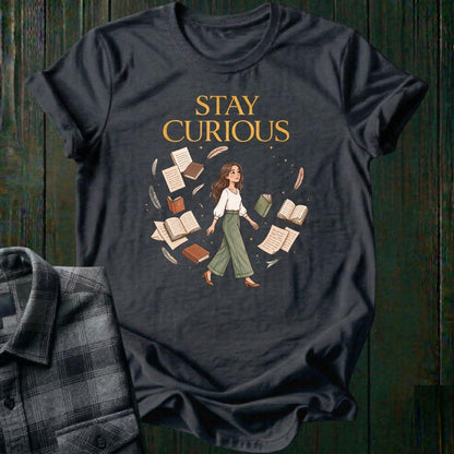 Illustrated woman walking among floating books, journals, feathers, and stars beneath the words "Stay Curious," symbolizing lifelong learning, creativity, and wonder on a charcoal tee.