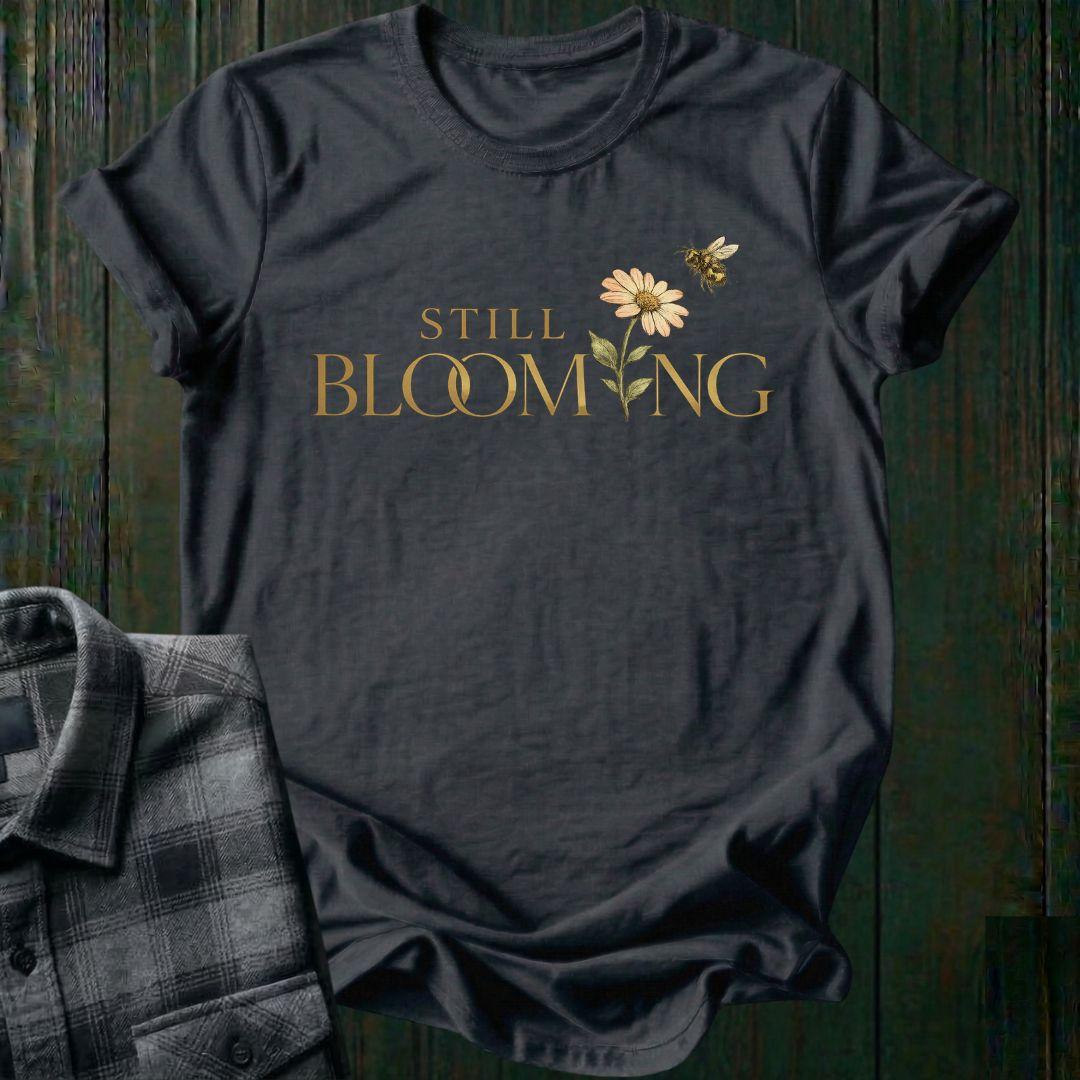 Elegant typography design featuring the phrase "Still Blooming" with a blooming flower growing through the lettering and a bee flying overhead on a charcoal shirt