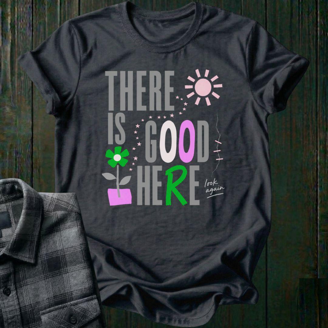 Colorful typography design featuring the phrase "There Is Good Here" with flowers, stars, sunshine, and the handwritten words "look again" on a charcoal tee.