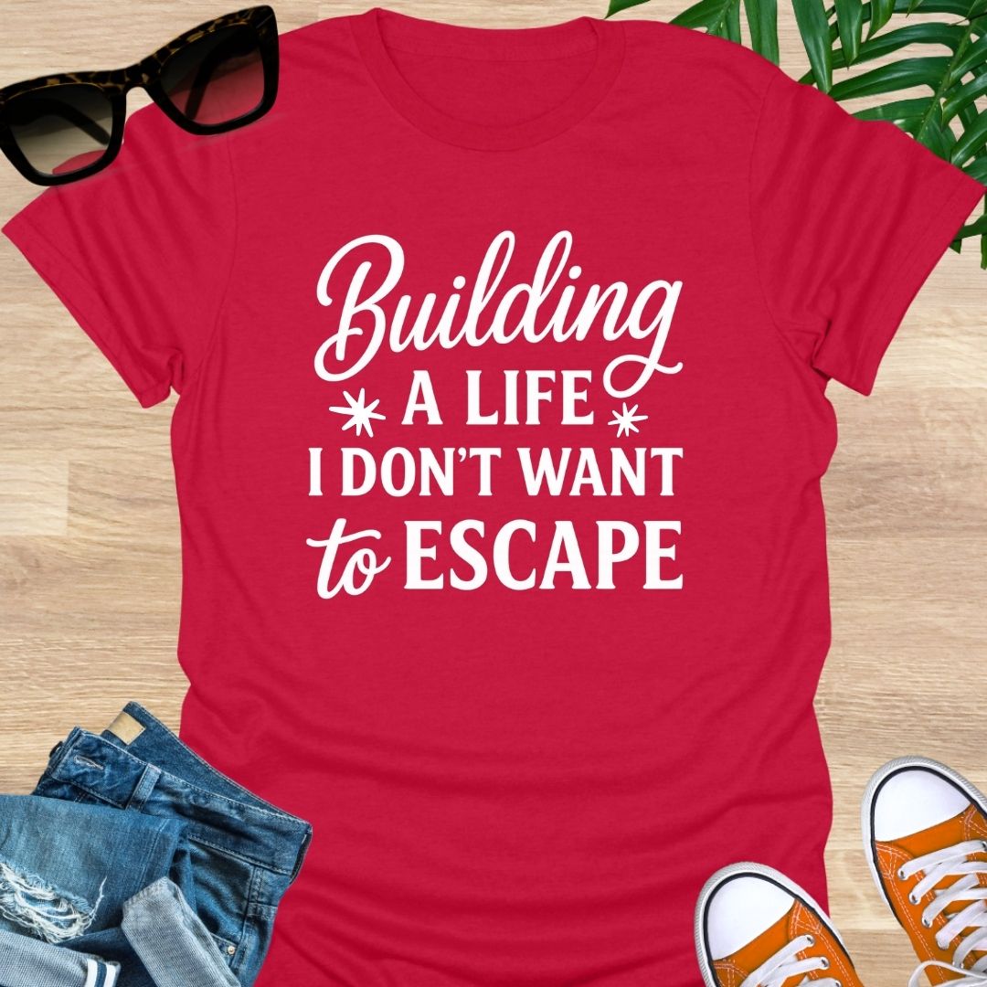 White script with block text: “Building a life I don’t want to escape” on red unisex tee.